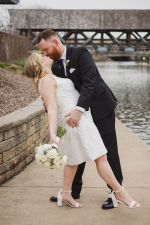 A bride and groom share a romantic dip kiss outdoors near a waterway, she holds white roses bouquet wearing short dress.