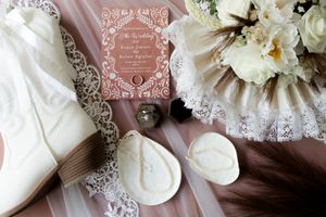 Wedding flat lay with lace veil, white boots, floral bouquet, rings, and elegant pink invitation card.
