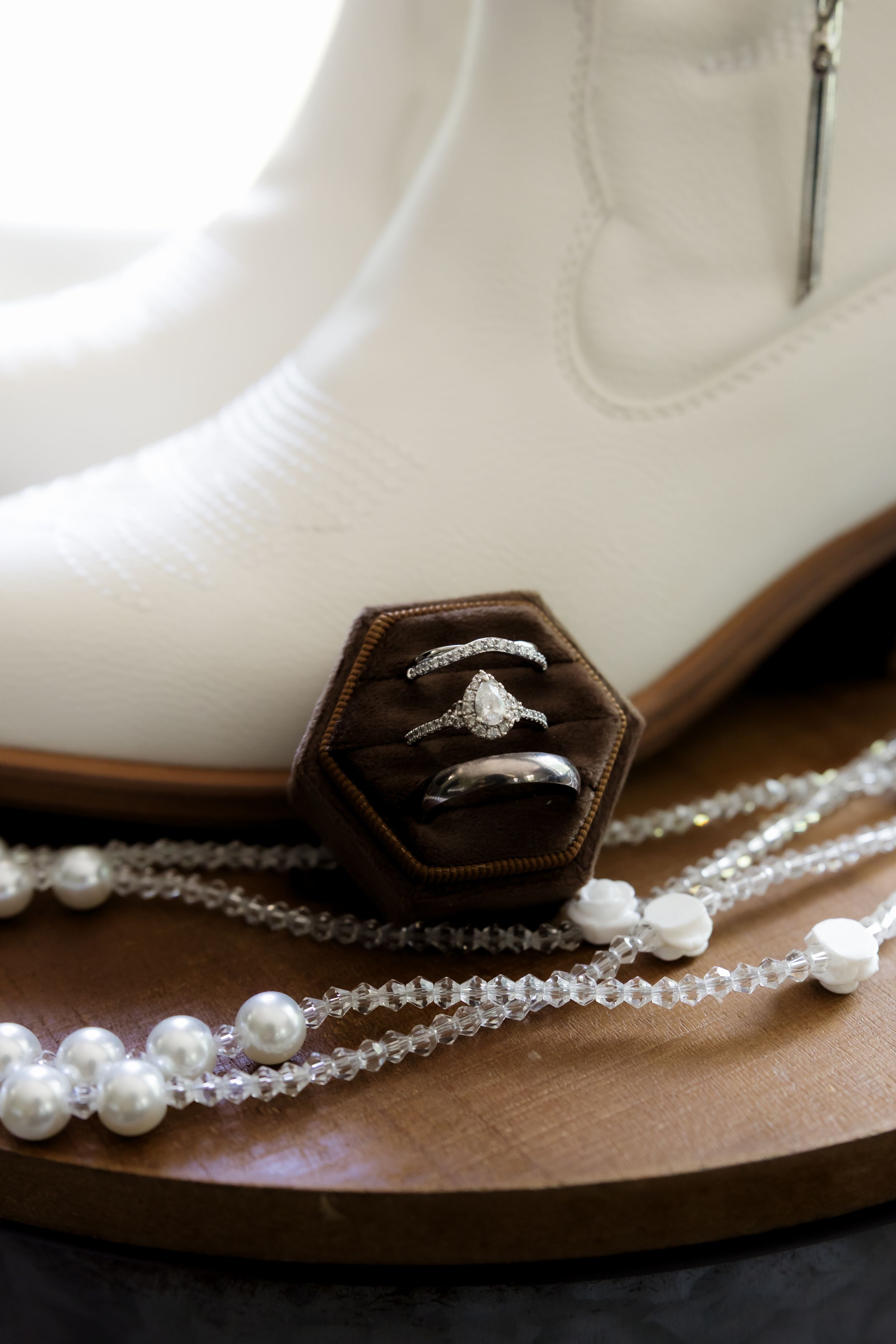 White cowboy boots with wedding rings in a brown velvet box, pearl and crystal necklaces on wooden surface.