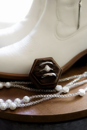 White cowboy boots with wedding rings in a brown velvet box, pearl and crystal necklaces on wooden surface.