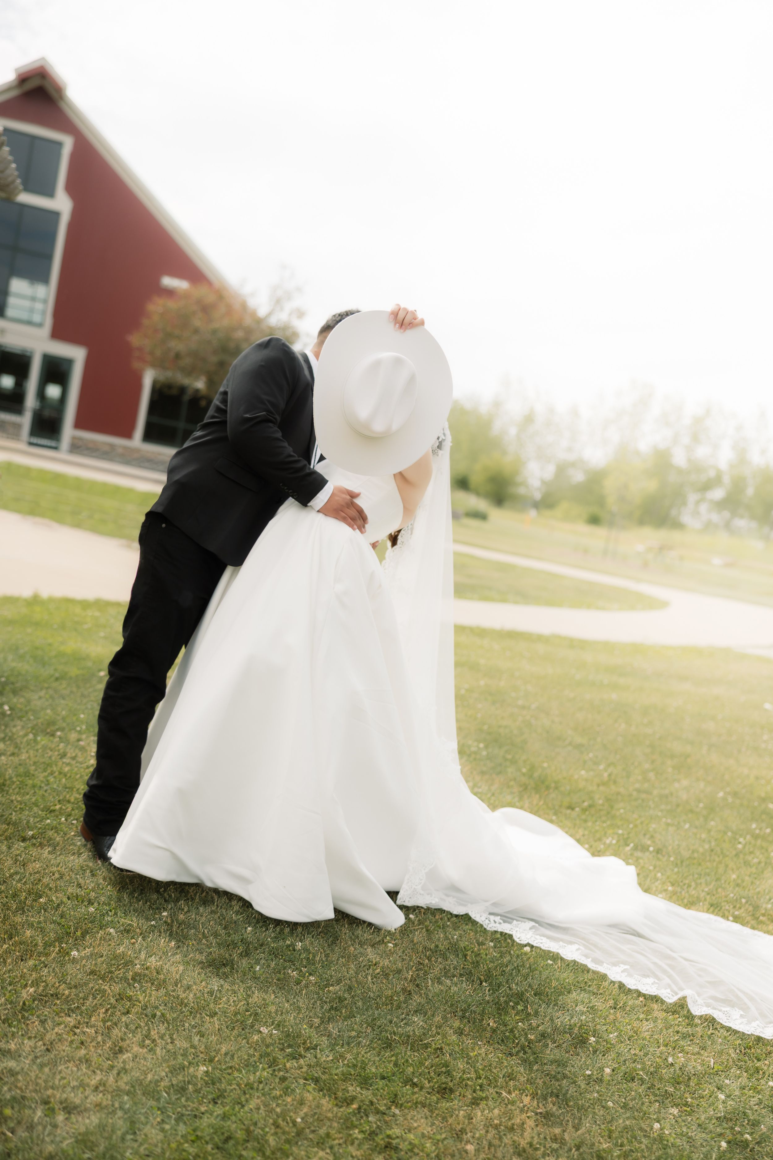 Bride and groom sharing a romantic kiss outdoors, bride wearing white gown and wide-brim hat, red barn in background.