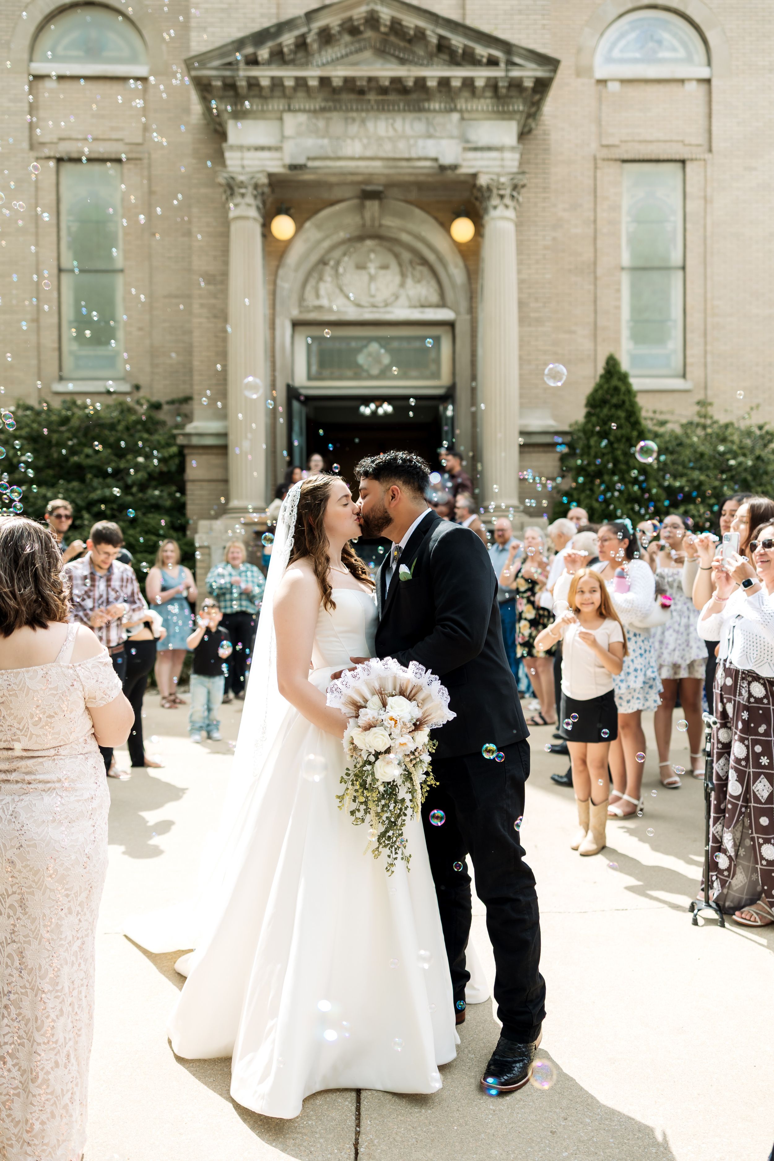 Newlywed couple sharing a kiss outside a historic church entrance, surrounded by cheering guests blowing bubbles.
