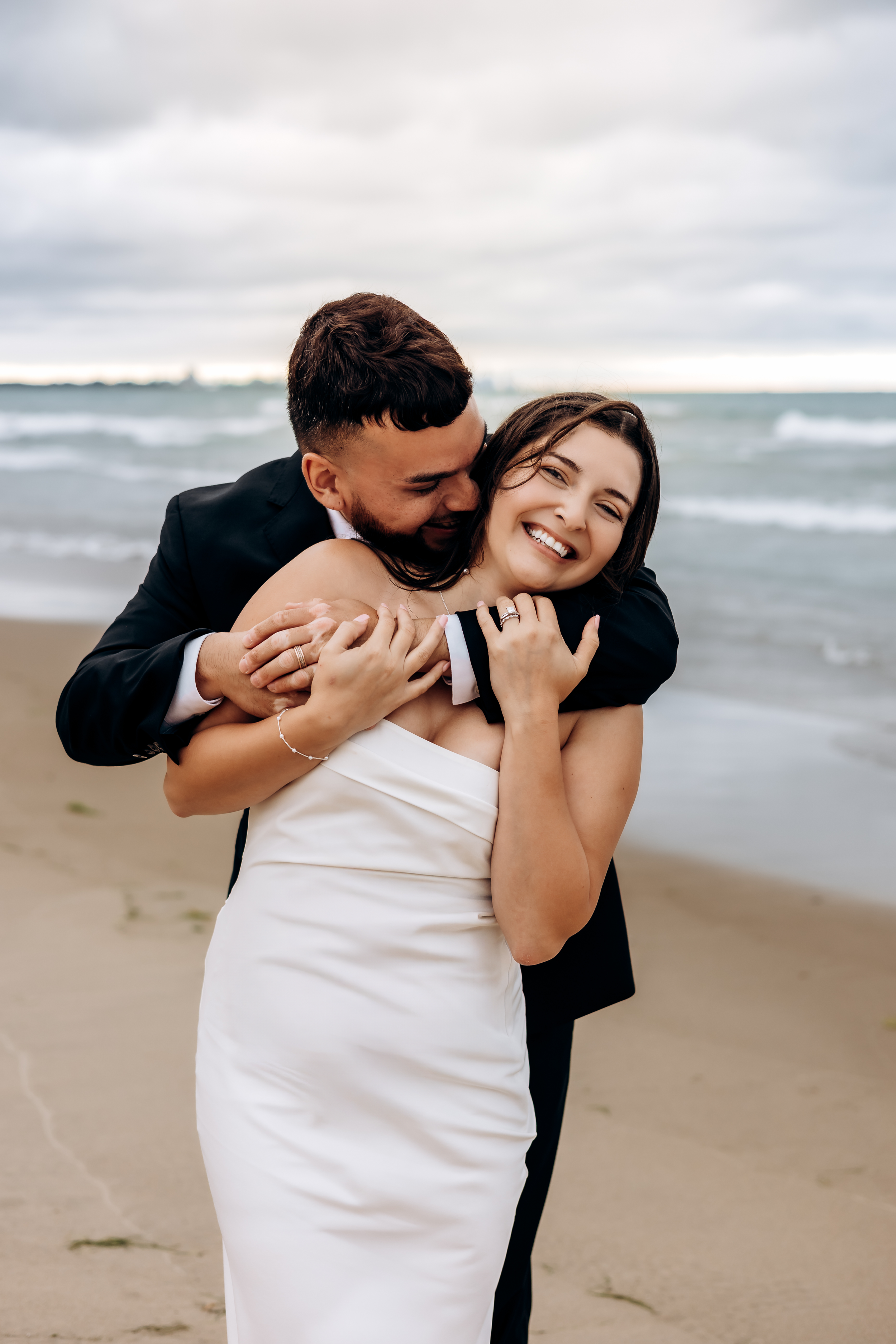 A joyful bride in a white dress embraces her groom in a black suit on a sandy beach with ocean waves behind them.