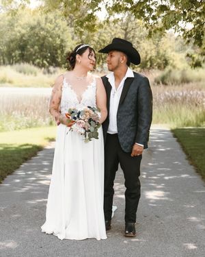 A bride in a white lace gown holds a bouquet beside a groom in a black cowboy hat and blazer on a garden path.