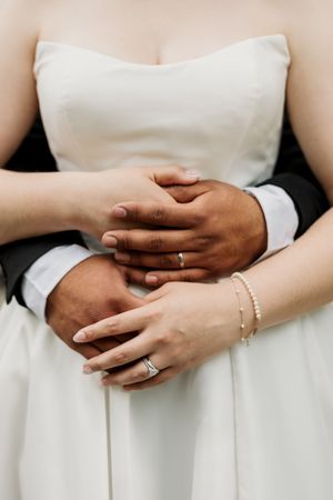 Close-up of interracial couple holding hands showing wedding rings, bride in white strapless gown with pearl bracelet.