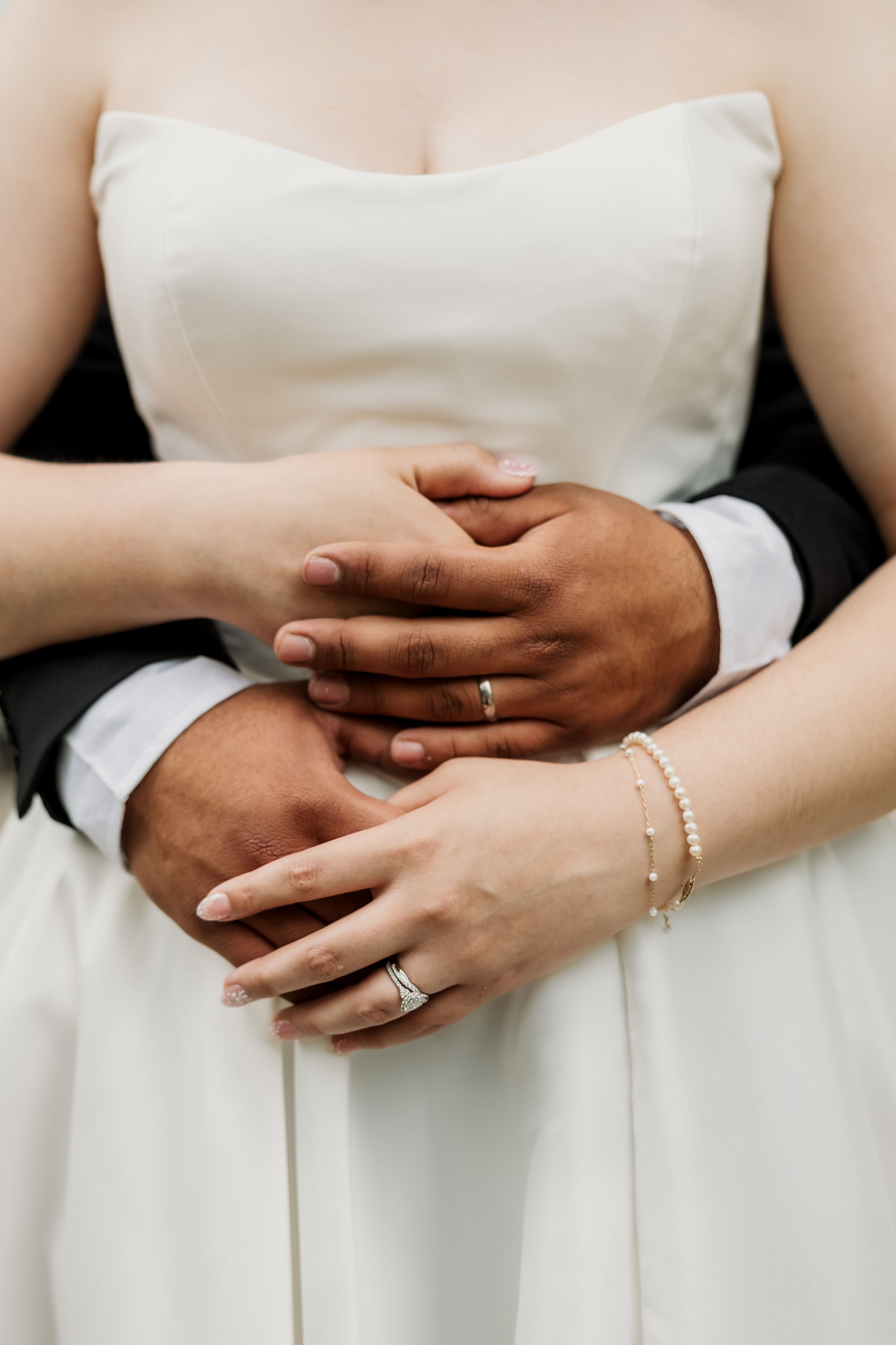 Close-up of interracial couple holding hands showing wedding rings, bride in white strapless gown with pearl bracelet.