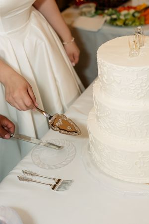 Bride cutting a three-tier white wedding cake with ornate silver cake server and knife set on white tablecloth.