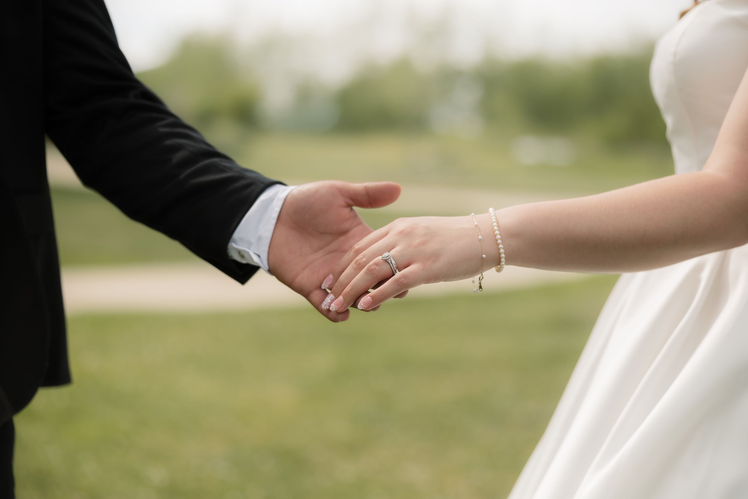 Bride and groom holding hands outdoors, bride wearing wedding ring and white dress, groom in black suit.