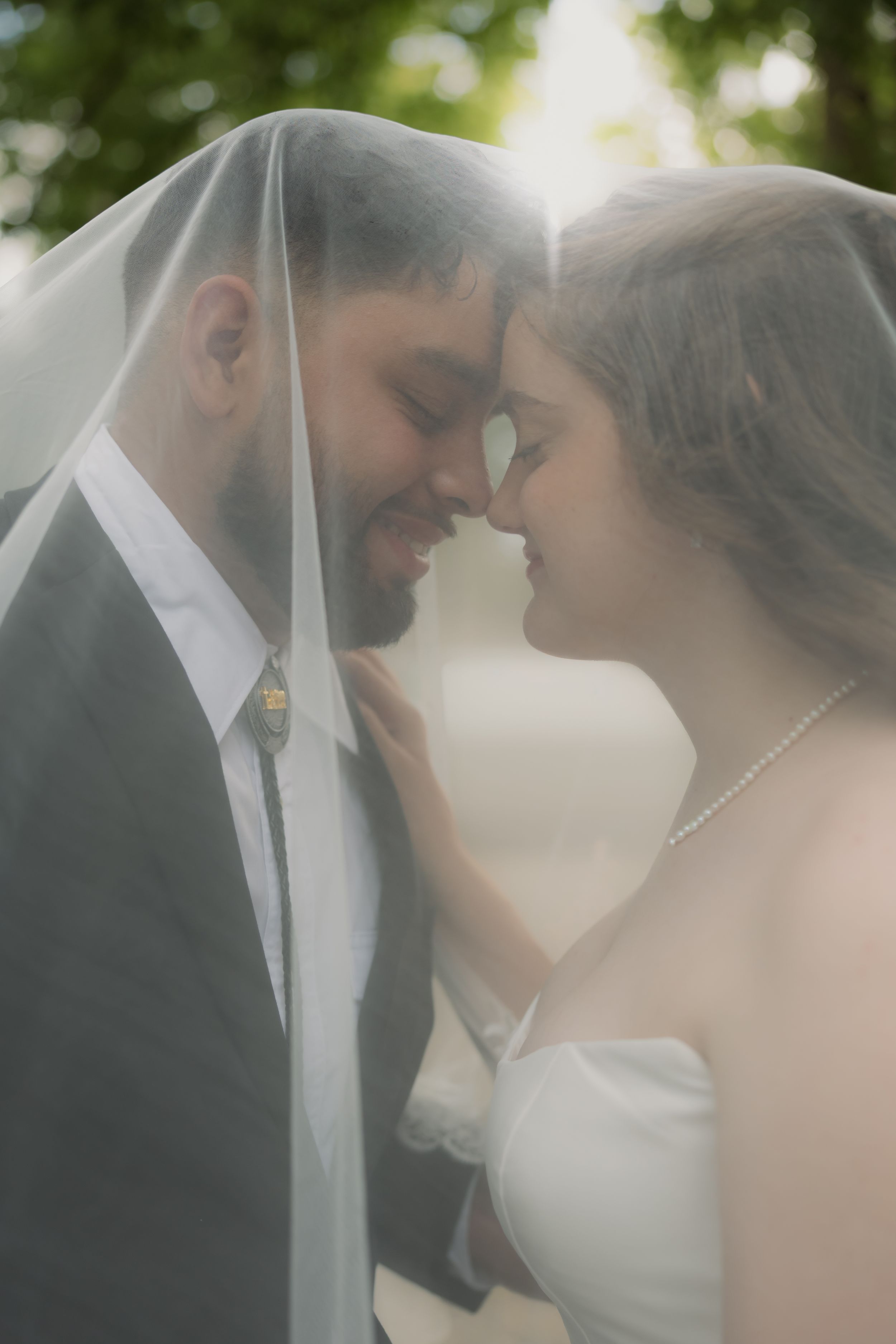 Bride and groom share an intimate moment under a wedding veil, touching foreheads and smiling softly.
