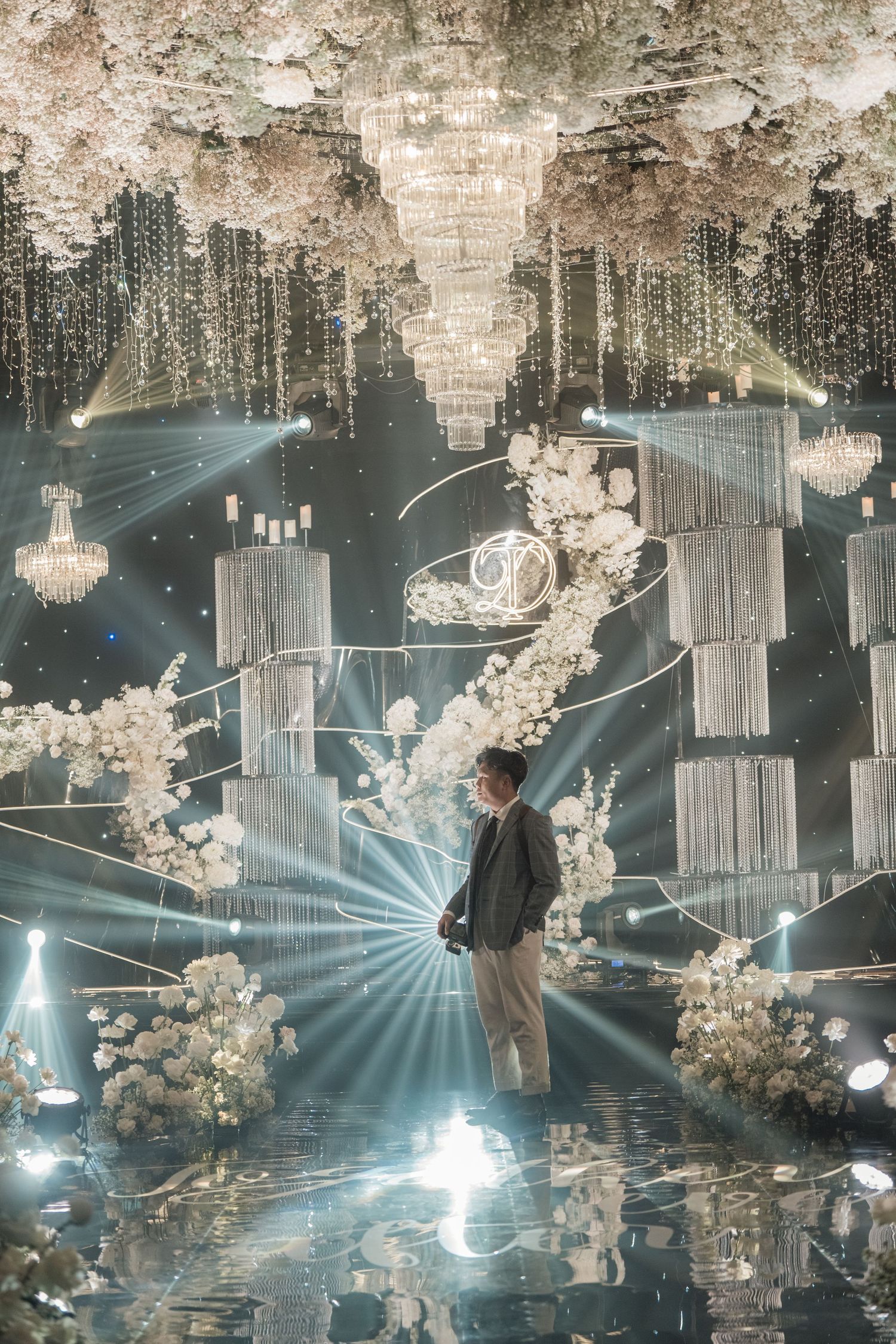 A man stands amid an elegant wedding venue with crystal chandeliers, white floral arrangements, and dramatic blue spotlight rays.