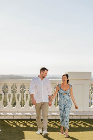 A smiling couple holds hands and walks on a sunlit terrace, man in white shirt and woman in blue floral jumpsuit.