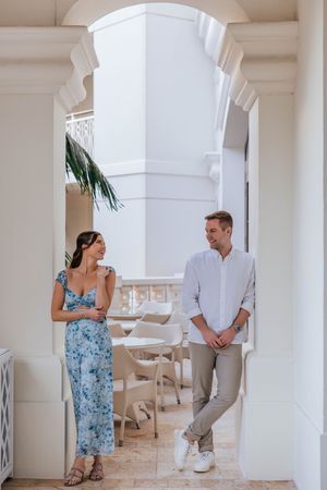 Couple conversing in elegant white archway, woman in blue floral dress, man in white shirt and khakis, resort setting.