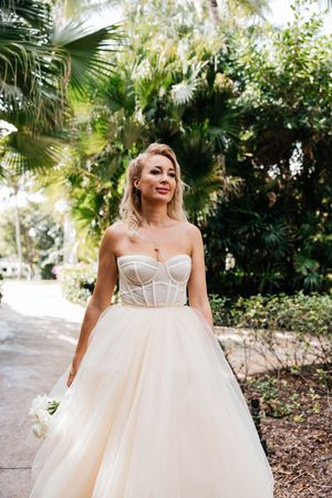 Elegant bride in strapless corset ball gown holding white bouquet among lush tropical greenery.