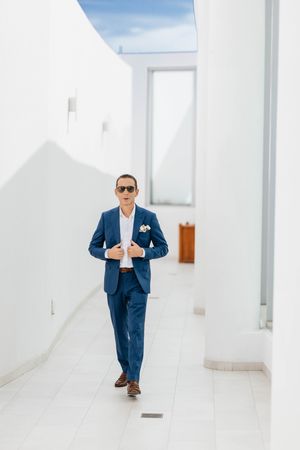 Stylish groom in blue suit and sunglasses walking through white corridor with boutonniere.