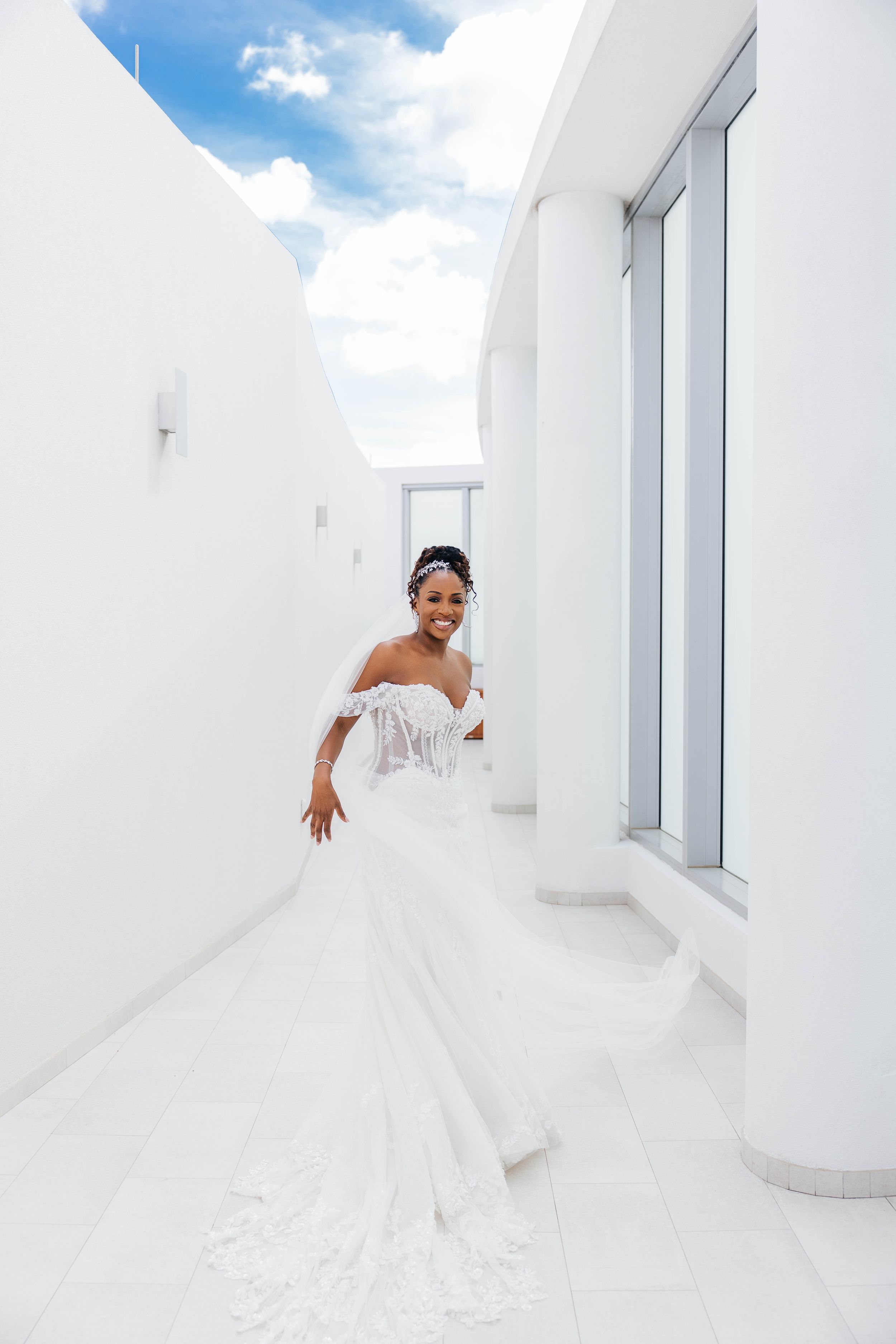 Smiling Black bride in off-shoulder white lace gown with long train poses in a bright white outdoor corridor.