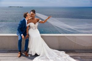 Interracial couple sharing a romantic kiss in wedding attire by the ocean, bride in white gown with veil.