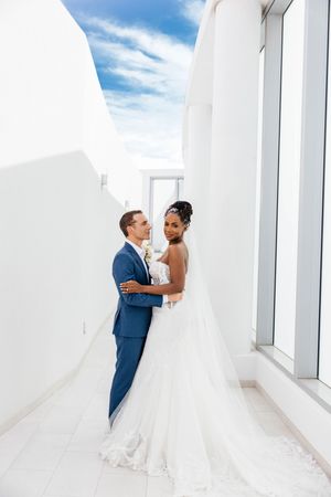 A bride and groom embrace in a white corridor, she in a flowing gown, he in a blue suit, under a bright blue sky.