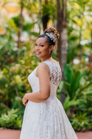 Smiling bride in lace wedding gown with floral hair accessory posing outdoors among tropical greenery.