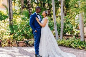 A bride and groom share a romantic moment in a lush tropical garden, with the groom in a blue suit and bride in a white gown.