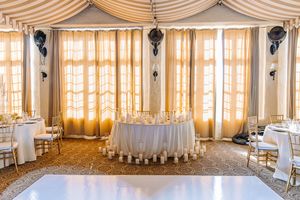 Elegant wedding reception venue with white draped sweetheart table, gold chairs, candles, and sheer curtains in a grand ballroom.