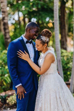 A joyful bride in a lace gown and groom in a blue tuxedo share an intimate moment outdoors amid tropical greenery.