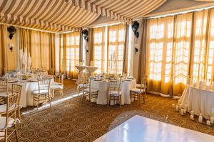 Elegant wedding reception hall with gold chiavari chairs, round tables, striped canopy ceiling, and warm draped windows.
