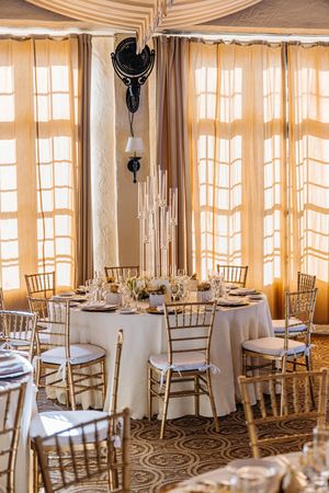 Luxurious wedding reception venue with gold chiavari chairs, white tablecloths, tall candelabras, and draped cream curtains.