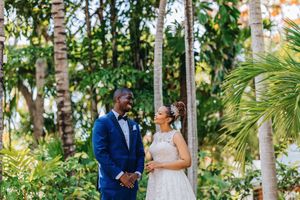 A bride and groom share a joyful moment surrounded by lush tropical palm trees in an outdoor wedding setting.