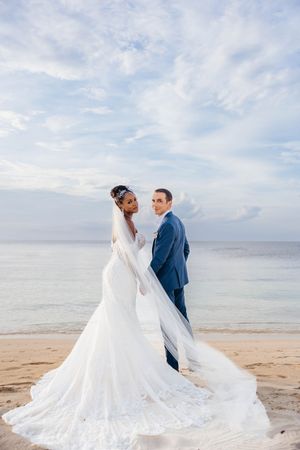Bride in white lace gown and groom in blue suit pose on sandy beach with ocean backdrop on their wedding day.
