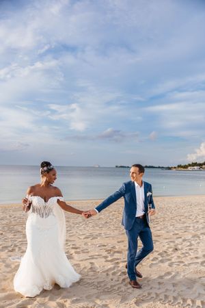 Bride and groom holding hands walking on sandy beach, wearing white gown and blue suit, with ocean backdrop.