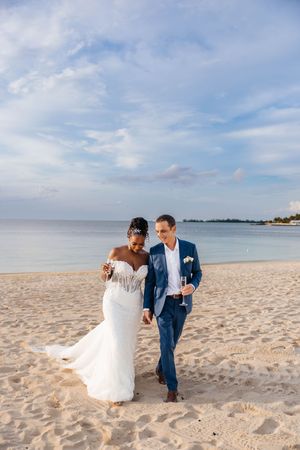 Bride and groom walking on sandy beach, woman in white mermaid gown, man in blue suit holding champagne.