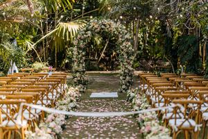 Romantic outdoor wedding ceremony aisle with floral arch, rose petal path, and wooden cross-back chairs in garden setting.
