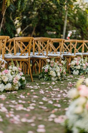 Outdoor wedding ceremony aisle with wooden cross-back chairs, pink and white floral arrangements, and rose petals scattered on grass.