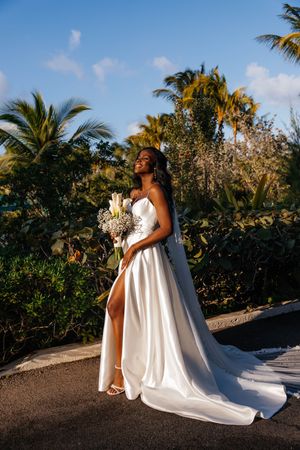 Radiant Black bride in white satin gown with thigh slit holding bouquet among lush tropical palm trees.