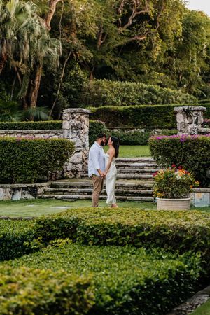Couple sharing a romantic moment in a lush garden with manicured hedges, stone steps, and tropical greenery.