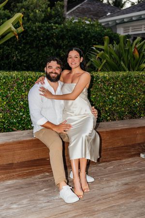 A smiling couple sits on a wooden bench outdoors, woman in silk dress and man in white shirt, embracing near tropical greenery.