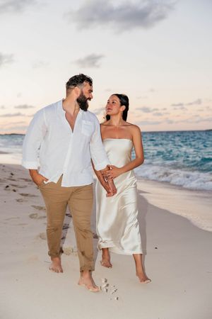 Couple in white attire walking barefoot on a sandy beach at sunset, holding hands near ocean waves.