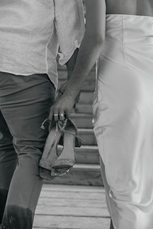 Black and white photo of couple holding hands, woman carrying heeled sandals on wooden steps.