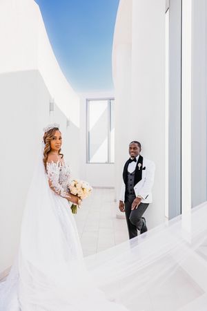 Bride in lace gown with tiara and groom in white tuxedo posing in a modern white architectural space.