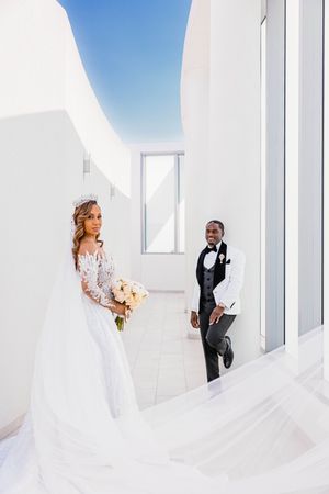 Bride in lace gown with crown bouquet and groom in tuxedo posing in a white architectural corridor.