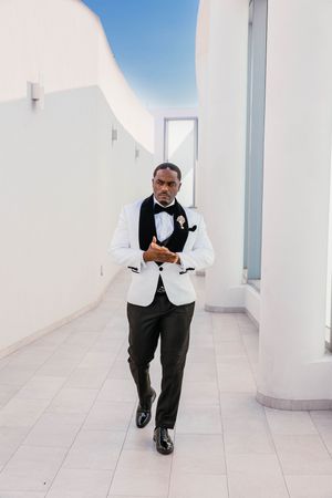 Stylish groom in white tuxedo jacket and black bow tie walking through a modern white corridor.