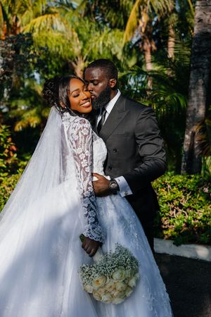 A groom in a black suit kisses his bride in a lace wedding gown holding a white bouquet outdoors among tropical palms.