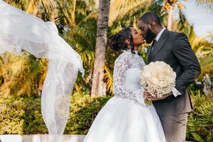 A bride and groom share a romantic kiss outdoors, with her white lace gown and flowing veil caught in the breeze.
