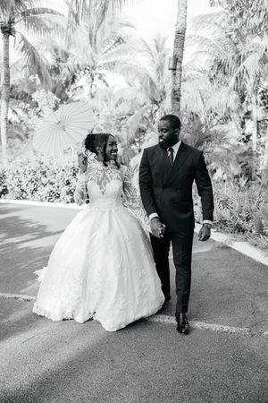 Black and white photo of a bride in a ball gown and groom in a suit walking hand in hand among palm trees.
