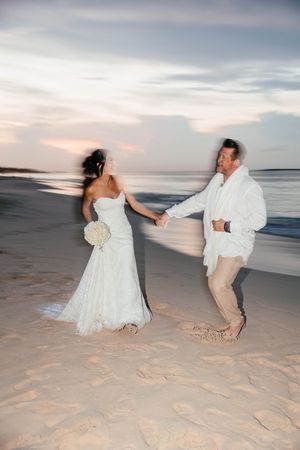 Joyful bride and groom holding hands and dancing on a sandy beach at sunset.