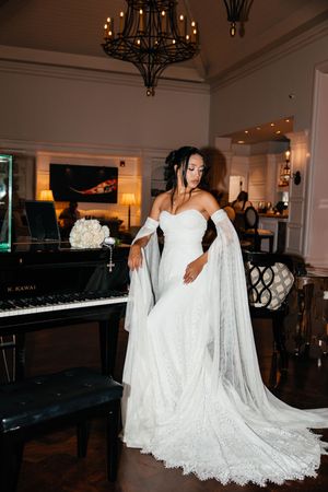 Bride in strapless white lace gown with flowing cape stands beside a black Kawai grand piano in elegant venue.