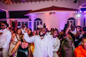 Joyful wedding reception guests dancing and celebrating in elegant white venue with purple ambient lighting.