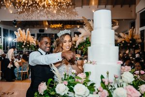 Bride and groom cutting elegant white tiered wedding cake surrounded by pink roses and pampas grass at reception.