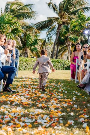 Young ring bearer runs down flower petal aisle at outdoor tropical wedding ceremony with palm trees.
