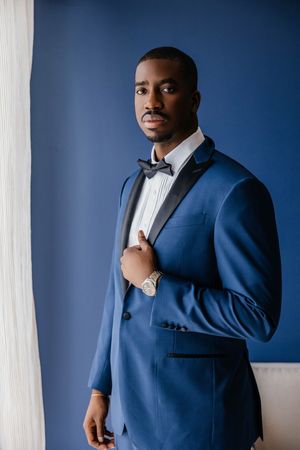 Elegant Black man in a blue tuxedo with bow tie and gold watch poses confidently against a navy blue wall.